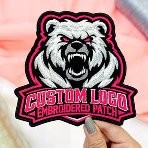 May include: An embroidered patch featuring a snarling white bear with glowing pink eyes and sharp teeth. The design includes a black border with pink accents and a banner at the bottom with the text CUSTOM LOGO EMBROIDERED PATCH in pink.