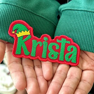 May include: A red embroidered patch with the name "Krista" in green, featuring a green elf hat with a gold crown. The patch is held against a green fabric background.