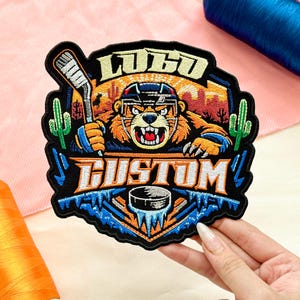 May include: Embroidered patch featuring a tiger wearing a hockey helmet and holding a hockey stick. The patch has the words "LOGO" and "CUSTOM" in a stylized font. The design includes a hockey puck, cacti, and a desert landscape. The patch is black with orange, blue, and white accents.