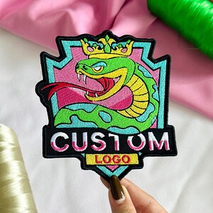 May include: An embroidered patch featuring a green snake wearing a yellow crown, set against a pink and light blue shield background. The black lower section displays the white text CUSTOM and the yellow text LOGO in a bold, block font.