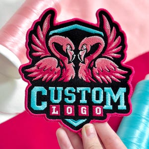 May include: Embroidered patch featuring two pink flamingos facing each other, with wings spread. The patch has a black background with the words "CUSTOM LOGO" in turquoise and white. The patch is surrounded by a pink border.