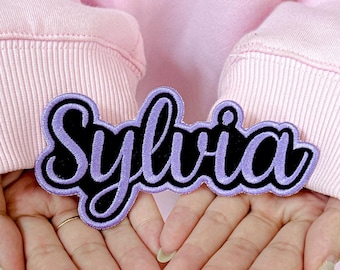May include: A custom embroidered patch in the shape of the name "Sylvia". The patch features a black base with the name in a cursive style, outlined in purple. The patch is held in front of a pink background.