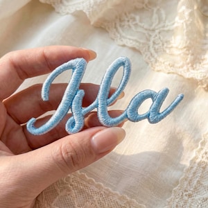 May include: A light blue embroidered patch spelling the name "Isla" in a cursive font. The patch is held in a hand, with the background featuring a soft, off-white lace fabric. The embroidery thread is a vibrant, sky blue.