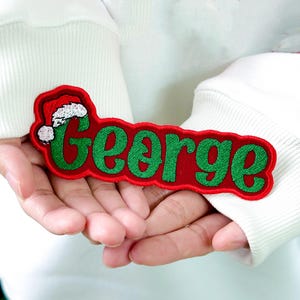 May include: A red embroidered patch with the name "George" in green script. The patch has a Santa hat design on the left side, perfect for Christmas. The patch is held in someone's hands.