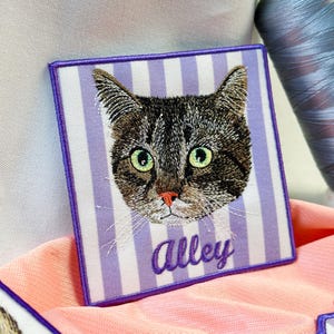 May include: A square embroidered patch featuring a brown tabby cat with green eyes and a pink nose. The background has vertical light purple and white stripes. The word Alley is embroidered in purple script below the cat's face.