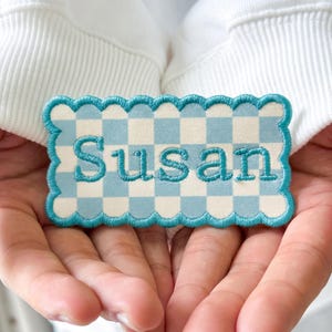 May include: A rectangular embroidered patch with a scalloped edge featuring a light blue and white checkered pattern. The name Susan is stitched in the center in a bold, dark teal thread, matching the border of the patch.