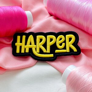 May include: A black patch embroidered with the name "HARPER" in yellow, outlined in black. The patch is set against a pink fabric background with several spools of pink thread.