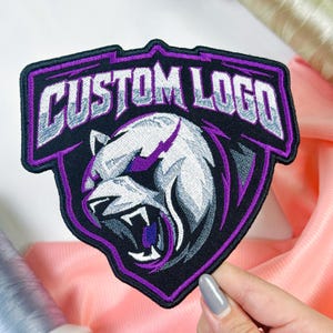 May include: A shield-shaped embroidered patch featuring a snarling panther head in shades of white, gray, and purple. The text CUSTOM LOGO is stitched in white with a gray gradient and a purple outline across the top of the black patch.