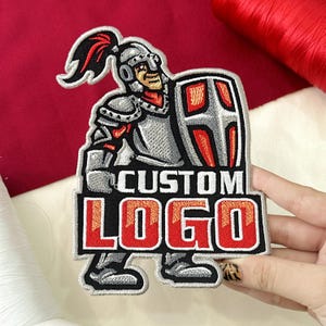May include: Embroidered patch featuring a knight in silver armor, holding a shield. The patch has the words "CUSTOM LOGO" in red and white. The knight has a red plume and is set against a red background.