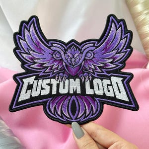 May include: An embroidered patch featuring a stylized purple owl with spread wings. The design includes a black border and the white text "CUSTOM LOGO" centered across the lower portion of the owl's body, set against a dark background.