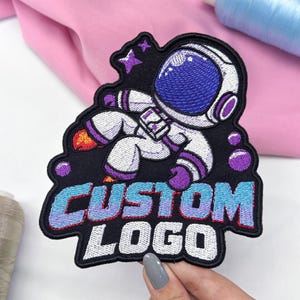 May include: An embroidered patch featuring an astronaut in a white and purple suit floating in space with stars and bubbles. The text CUSTOM LOGO is stitched in a blue to purple gradient font at the bottom. The patch has a black border.