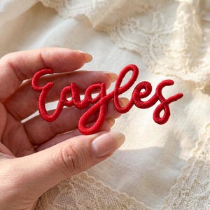 May include: A red embroidered patch with the word "Eagles" in a cursive script. The patch is made of a thick, textured thread and is likely designed to be sewn onto clothing or accessories. The patch is held in a hand.