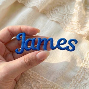 May include: A blue embroidered patch with the name "James" in a cursive font. The patch is held in a hand against a background of white lace fabric.