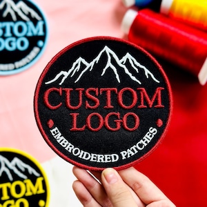 May include: Embroidered patch with a black background, red border, and white mountain range design. The words "CUSTOM LOGO" are in red, and "EMBROIDERED PATCHES" are in white. The patch is round and held by a hand.