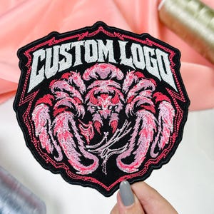 May include: A black embroidered patch featuring a central pink and white spider design with the text CUSTOM LOGO in white block letters arched across the top. The patch has a scalloped black border with a thin pink stitched outline.