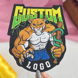 May include: A black embroidered patch featuring a muscular orange panther character with blue eyes, wearing blue shorts and holding a brown wooden club wrapped in a green snake. The word CUSTOM is in yellow and green above, and LOGO is in white below.
