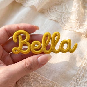 May include: A mustard yellow embroidered patch featuring the name Bella in a cursive script font. The lettering is formed by thick, raised thread work with a satin finish, creating a dimensional appearance for the custom name design.