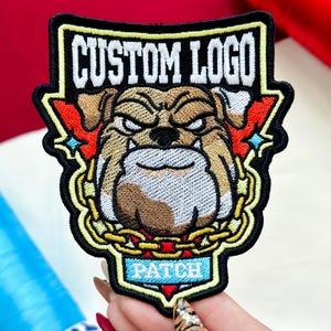 May include: Embroidered patch featuring a bulldog with an angry expression, surrounded by a gold chain and red accents. The patch has the words "CUSTOM LOGO" and "PATCH" in white and blue, respectively, on a black background.