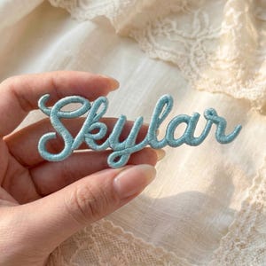 May include: A light blue embroidered patch with the name "Skylar" in cursive script. The patch is held in a hand against a soft, textured background. The embroidery thread is a soft, pastel blue.