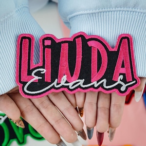May include: A rectangular embroidered patch with the name "LINDA" in pink, outlined in black, on a black background. The last name "EVANS" is embroidered in white cursive below. The patch is held against a light blue background.