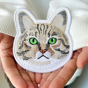 May include: Embroidered cat face patch with green eyes, tan and brown fur, and a white border. The patch is held in front of a white background. This patch is perfect for adding to clothing or accessories.