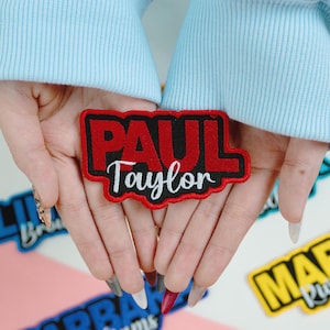 May include: A red and black embroidered patch with the name "PAUL Taylor" in white script. The patch is rectangular with a black border and is held in open hands. Other patches are visible in the background.