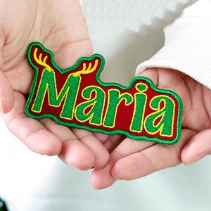 May include: A red and green embroidered patch with the name "Maria" in green, outlined in yellow. The patch has a festive design with antlers, perfect for personalization or holiday crafts.
