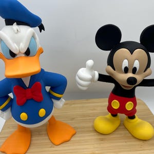 Op de afbeelding: Twee kleurrijke beeldjes van Disney-personages: Donald Duck en Mickey Mouse. Donald Duck draagt een blauw matrozenpak met een rode vlinderdas. Mickey Mouse draagt een rode broek en gele schoenen, met zijn armen omhoog in een duim omhoog gebaar.