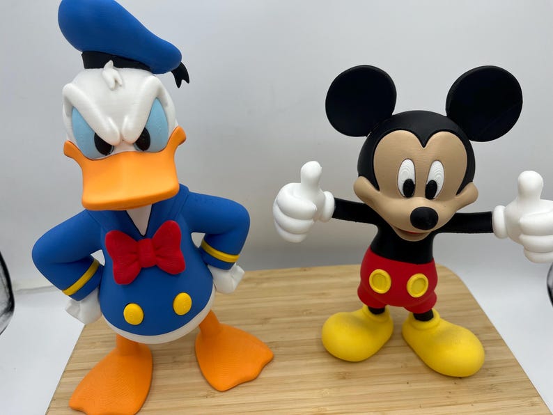 Op de afbeelding: Twee kleurrijke beeldjes van Disney-personages: Donald Duck en Mickey Mouse. Donald Duck draagt een blauw matrozenpak met een rode vlinderdas. Mickey Mouse draagt een rode broek en gele schoenen, met zijn armen omhoog in een duim omhoog gebaar.