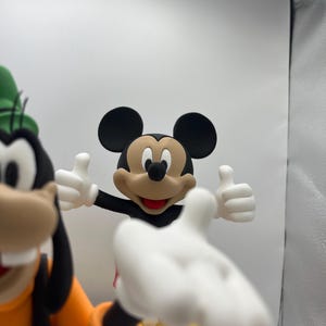 Op de afbeelding: Een Mickey Mouse-beeldje met uitgestrekte armen, die een duim omhoog geeft. Mickey Mouse draagt een zwarte broek, een rood shirt en gele schoenen. Goofy staat op de achtergrond en draagt een oranje shirt en een groene hoed.