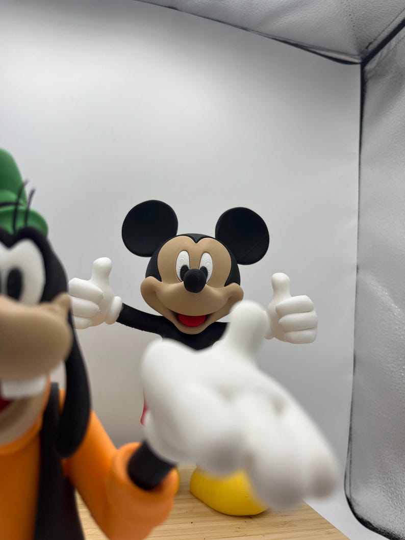 Op de afbeelding: Een Mickey Mouse-beeldje met uitgestrekte armen, die een duim omhoog geeft. Mickey Mouse draagt een zwarte broek, een rood shirt en gele schoenen. Goofy staat op de achtergrond en draagt een oranje shirt en een groene hoed.