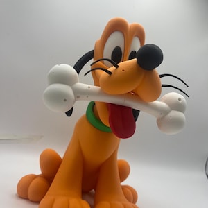 Op de afbeelding: Een beeldje van Pluto, een cartoonhond, in een zittende positie. De oranje hond heeft een zwarte neus, oren en wenkbrauwen en een rode tong. Hij houdt een wit bot in zijn mond en draagt een groene halsband. Het beeldje staat op een witte achtergrond.
