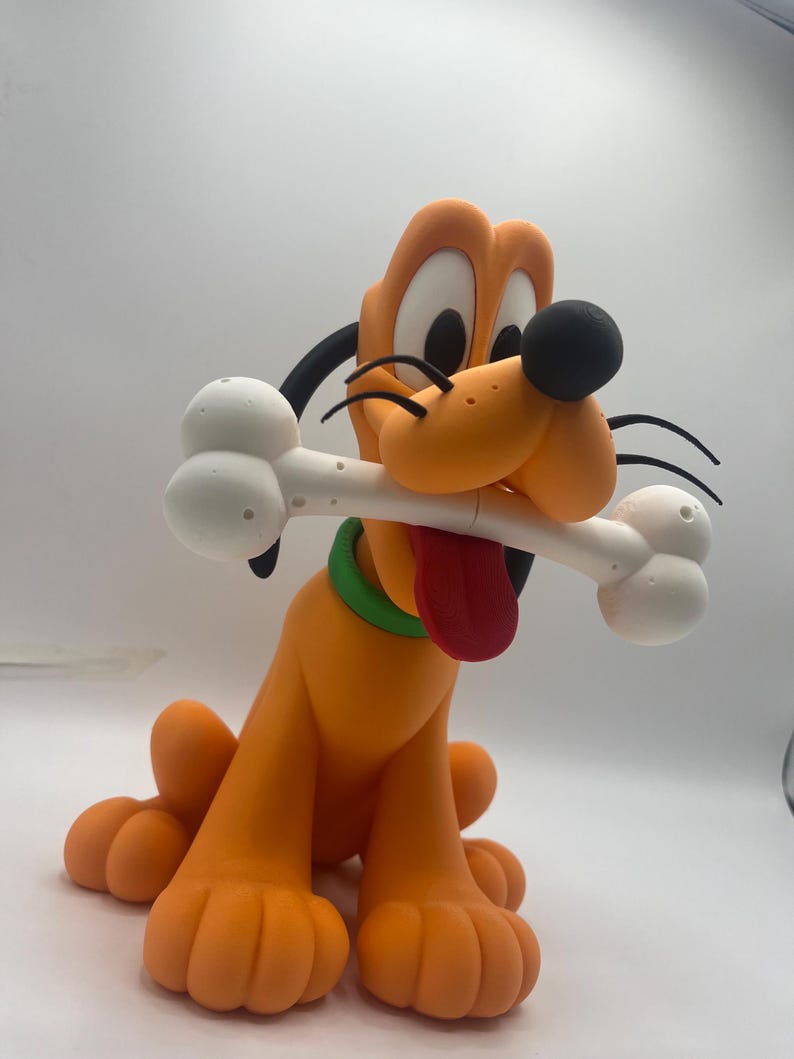 Op de afbeelding: Een beeldje van Pluto, een cartoonhond, in een zittende positie. De oranje hond heeft een zwarte neus, oren en wenkbrauwen en een rode tong. Hij houdt een wit bot in zijn mond en draagt een groene halsband. Het beeldje staat op een witte achtergrond.