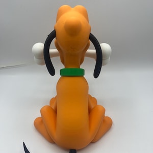 Op de afbeelding: Oranje beeldje van een cartoonhond, van achteren gezien, met een wit bot in zijn bek. De hond heeft zwarte oren, een groene kraag en een zwarte staart. Het beeldje staat tegen een effen witte achtergrond.