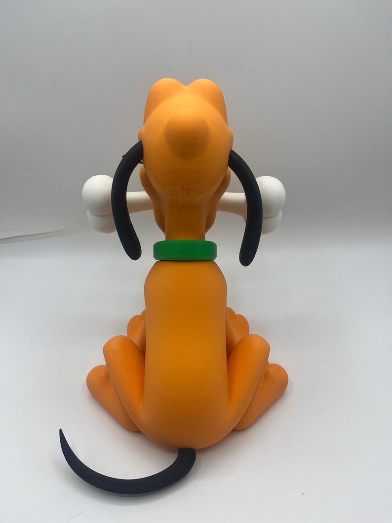 Op de afbeelding: Oranje beeldje van een cartoonhond, van achteren gezien, met een wit bot in zijn bek. De hond heeft zwarte oren, een groene kraag en een zwarte staart. Het beeldje staat tegen een effen witte achtergrond.