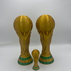 Puede incluir: Tres trofeos dorados de la Copa Mundial de la FIFA de diferentes tamaños. Los dos trofeos más grandes tienen un diseño de globo en la parte superior y una base verde con el texto "FIFA WORLD CUP". El trofeo más pequeño tiene un diseño similar.