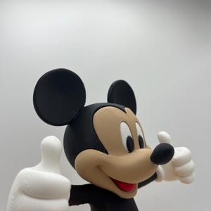 Op de afbeelding: Een 3D-geprinte figuur van Mickey Mouse, een klassiek stripfiguur, met een zwarte kop en oren, een bruin gezicht en een rode en zwarte outfit. De figuur geeft een duim omhoog met beide witte handschoenen.