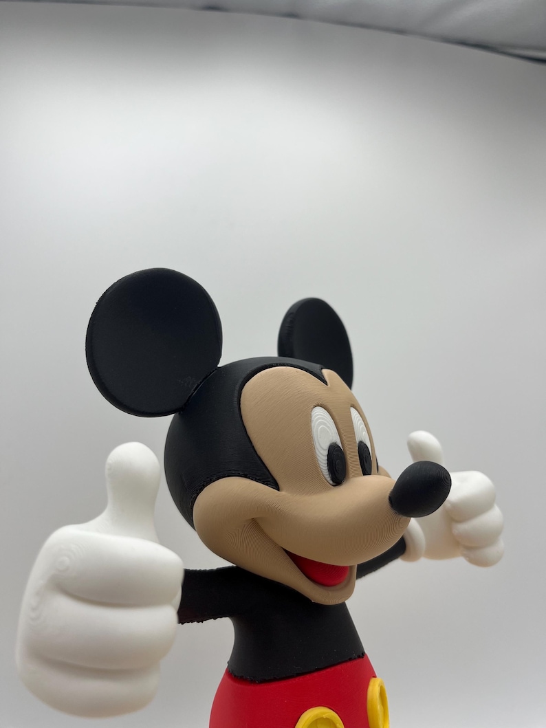 Op de afbeelding: Een 3D-geprinte figuur van Mickey Mouse, een klassiek stripfiguur, met een zwarte kop en oren, een bruin gezicht en een rode en zwarte outfit. De figuur geeft een duim omhoog met beide witte handschoenen.