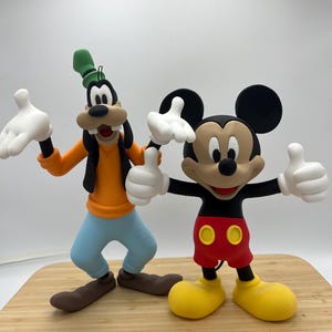 Op de afbeelding: Twee stripfiguurbeeldjes, Goofy en Mickey Mouse, staan naast elkaar. Goofy draagt een oranje shirt, een blauwe broek en een groene hoed. Mickey Mouse draagt een rode korte broek, gele schoenen en zwarte oren. Beide personages hebben witte handschoenen en lachen.