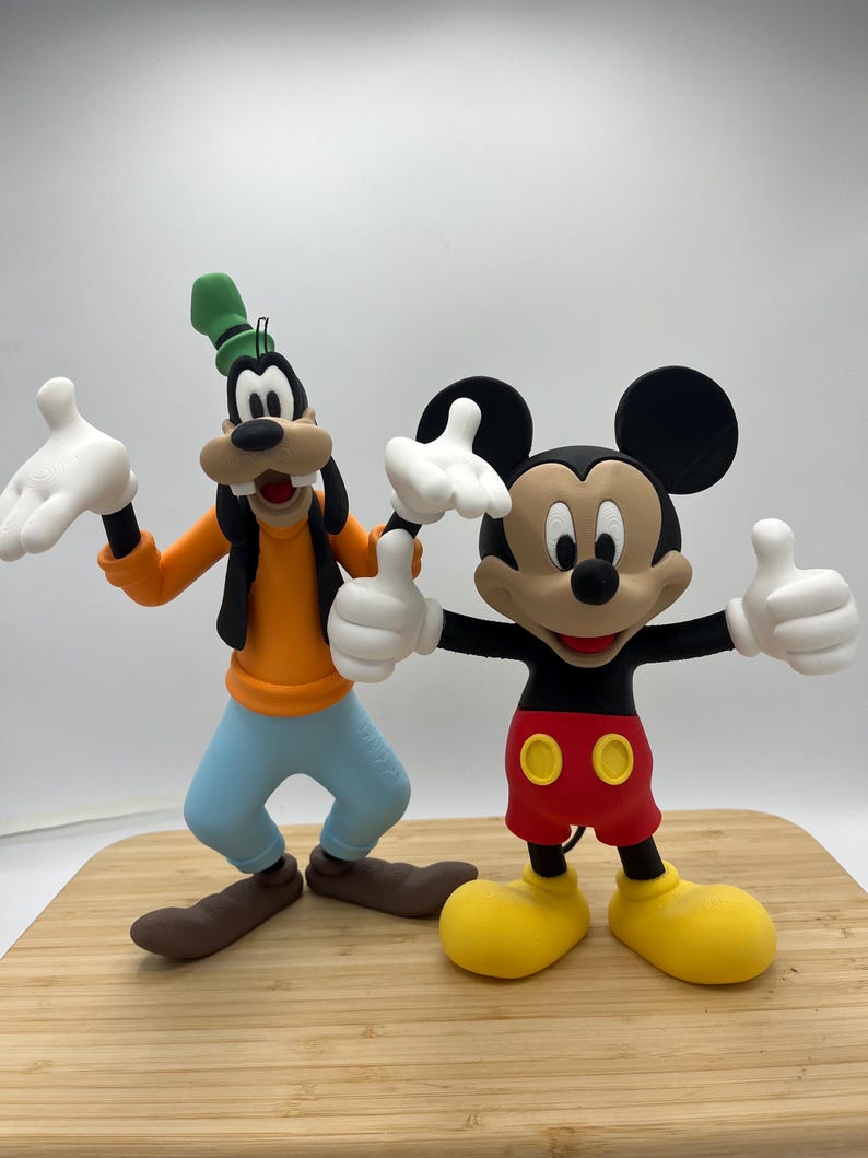 Op de afbeelding: Twee stripfiguurbeeldjes, Goofy en Mickey Mouse, staan naast elkaar. Goofy draagt een oranje shirt, een blauwe broek en een groene hoed. Mickey Mouse draagt een rode korte broek, gele schoenen en zwarte oren. Beide personages hebben witte handschoenen en lachen.