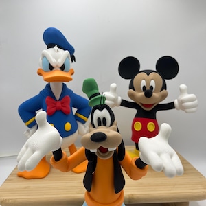Op de afbeelding: Drie Disney-personagefiguren: Donald Duck in een blauw matrozenpak, Mickey Mouse in een rode broek en Goofy in een oranje shirt en blauwe broek. De figuren staan op een houten oppervlak met een witte achtergrond.