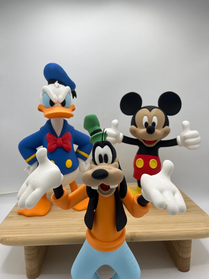 Op de afbeelding: Drie Disney-personagefiguren: Donald Duck in een blauw matrozenpak, Mickey Mouse in een rode broek en Goofy in een oranje shirt en blauwe broek. De figuren staan op een houten oppervlak met een witte achtergrond.