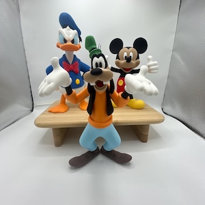 Op de afbeelding: Drie stripfiguurbeeldjes: Goofy in oranje en blauw, Mickey Mouse in rood en zwart en Donald Duck in blauw. Ze staan op een kleine houten tafel tegen een witte achtergrond. De karakters staan in een speelse pose.