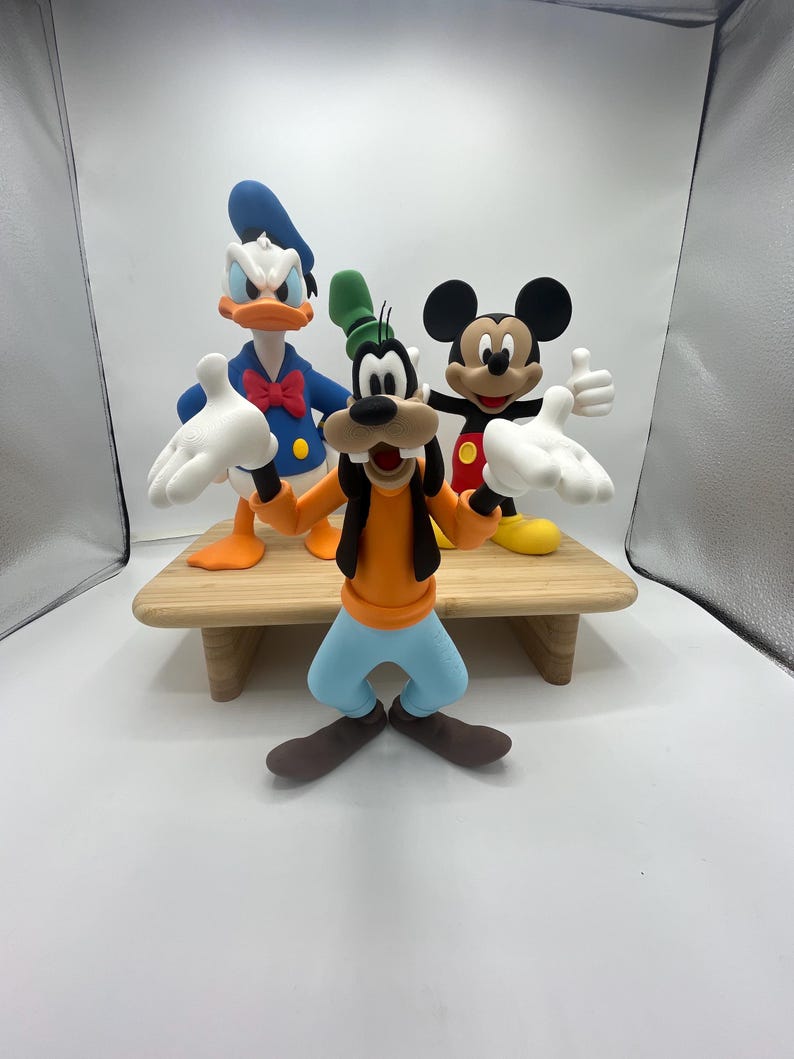 Op de afbeelding: Drie stripfiguurbeeldjes: Goofy in oranje en blauw, Mickey Mouse in rood en zwart en Donald Duck in blauw. Ze staan op een kleine houten tafel tegen een witte achtergrond. De karakters staan in een speelse pose.