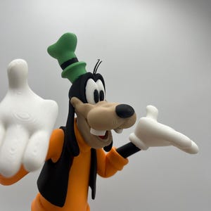 Op de afbeelding: Een 3D-geprinte figuur van Goofy, een Disney-stripfiguur. Goofy draagt een oranje shirt, een zwart vest en een blauwe broek. Zijn hoed is groen met een zwarte band en hij heeft witte handschoenen. De figuur staat tegen een effen achtergrond.