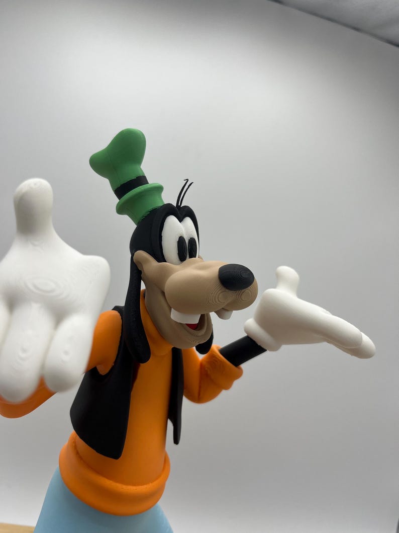 Op de afbeelding: Een 3D-geprinte figuur van Goofy, een Disney-stripfiguur. Goofy draagt een oranje shirt, een zwart vest en een blauwe broek. Zijn hoed is groen met een zwarte band en hij heeft witte handschoenen. De figuur staat tegen een effen achtergrond.