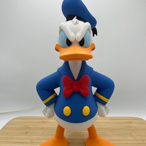 Op de afbeelding: Een beeldje van Donald Duck, een stripfiguur, met een boze uitdrukking. De figuur is voornamelijk blauw, wit en oranje, met een rode vlinderdas en een blauwe matrozenpet. Het heeft witte handschoenen en oranje voeten.