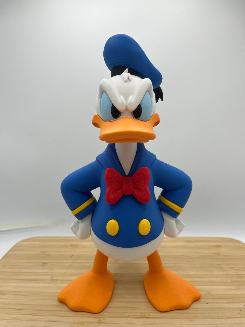 Op de afbeelding: Een beeldje van Donald Duck, een stripfiguur, met een boze uitdrukking. De figuur is voornamelijk blauw, wit en oranje, met een rode vlinderdas en een blauwe matrozenpet. Het heeft witte handschoenen en oranje voeten.