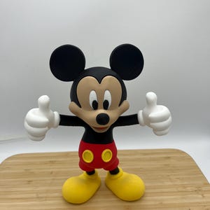 Op de afbeelding: Een Mickey Mouse-beeldje met zwarte oren, een zwarte neus en een lachend gezicht. Hij draagt een rode broek, gele schoenen en witte handschoenen. Het beeldje staat op een houten oppervlak, met beide duimen omhoog.