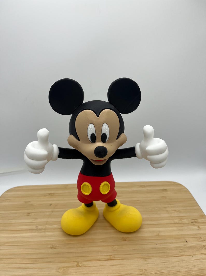 Op de afbeelding: Een Mickey Mouse-beeldje met zwarte oren, een zwarte neus en een lachend gezicht. Hij draagt een rode broek, gele schoenen en witte handschoenen. Het beeldje staat op een houten oppervlak, met beide duimen omhoog.